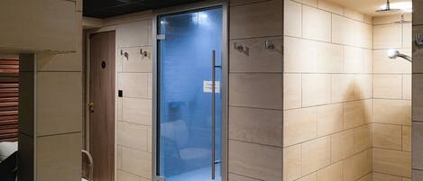 A modern bathroom with a glass door, beige tiles, a shower area, and a seating area with a plant.