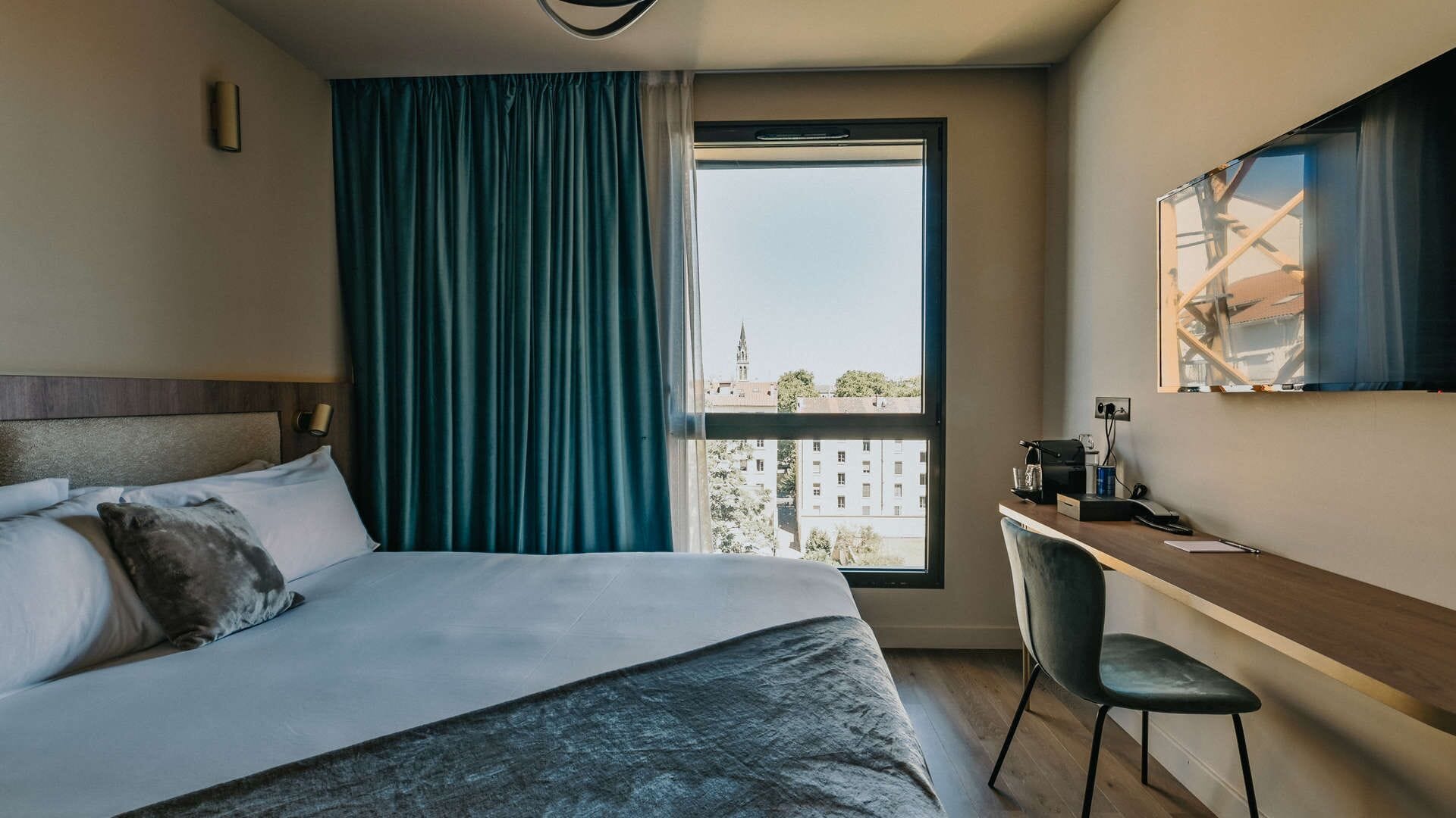 Camera d'albergo con un letto, una scrivania, una sedia, una finestra vista edifici e una tenda.