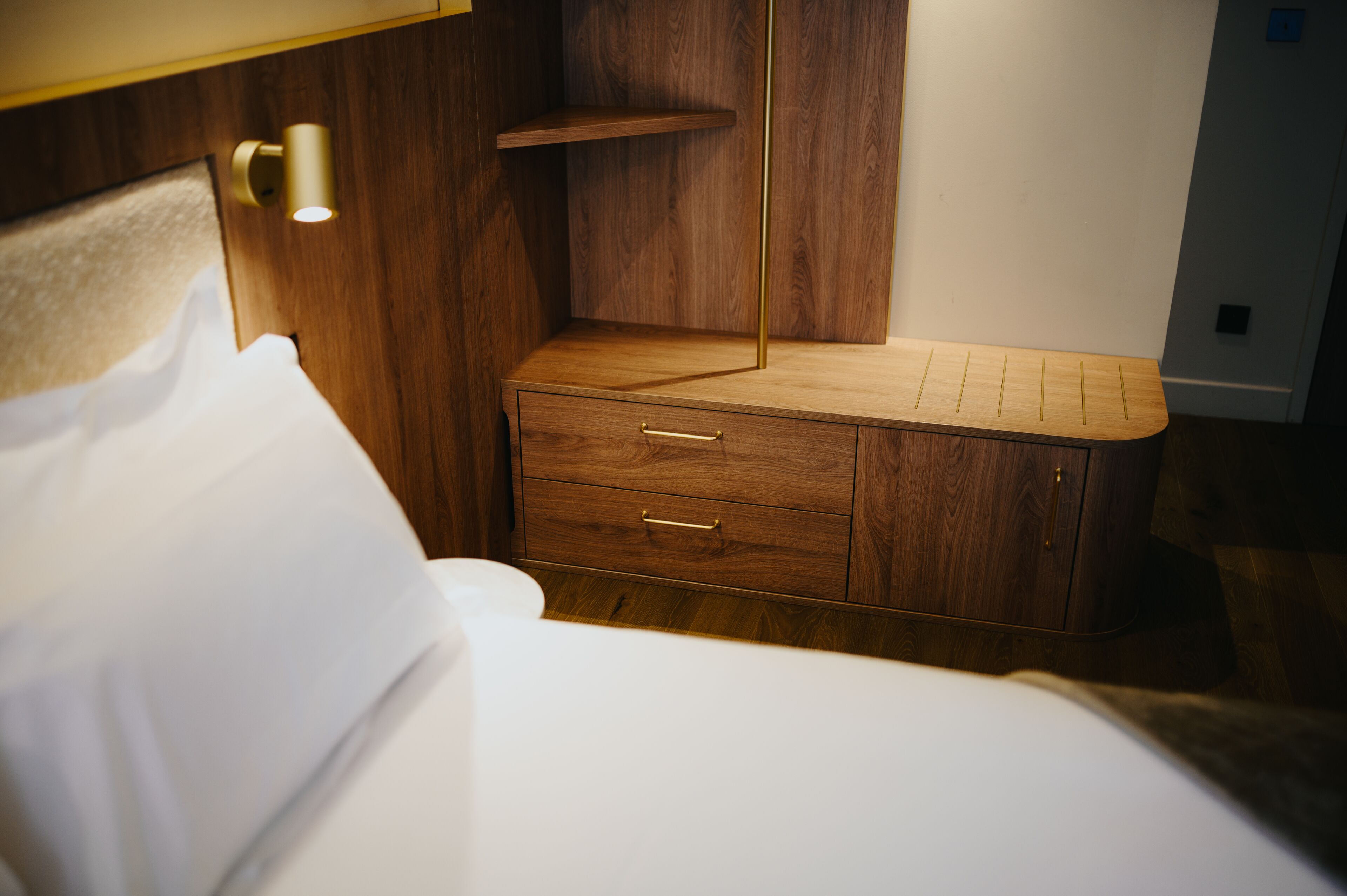 A wooden bedside table with drawers, a nightstand, and a wall-mounted shelf in a hotel room.