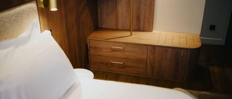 A wooden bedside table with drawers, a nightstand, and a wall-mounted shelf in a hotel room.