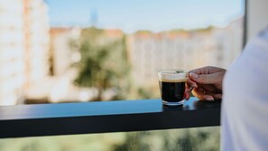 A hand holding a glass of dark liquid, possibly coffee, on a balcony with a blurred cityscape in the background. - Keystone Hotel & Spa Lyon - Handwritten Collection (Lyon)