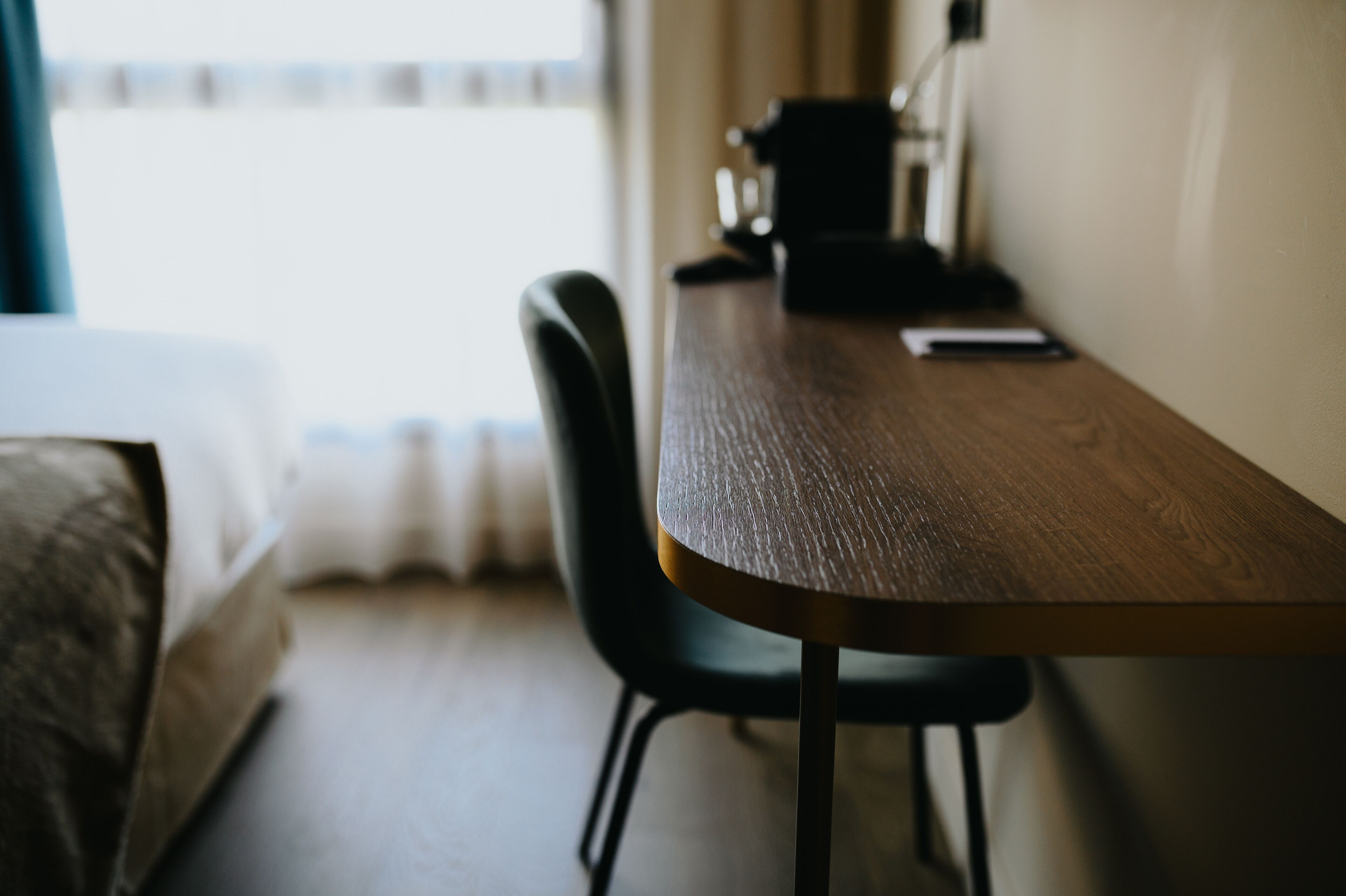 A room with a wooden table, a black chair, a bed, and a window with curtains.