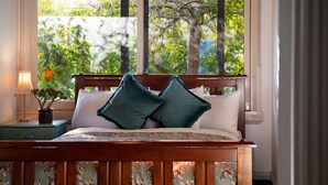 Un lit avec une tête de lit en bois, deux oreillers, une table de chevet avec une lampe et une plante, et une fenêtre donnant sur des arbres.