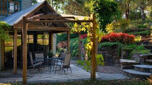 Une pergola en bois avec une table et des chaises, entourée d’un jardin où poussent différentes plantes et bordée d’un mur en pierre.