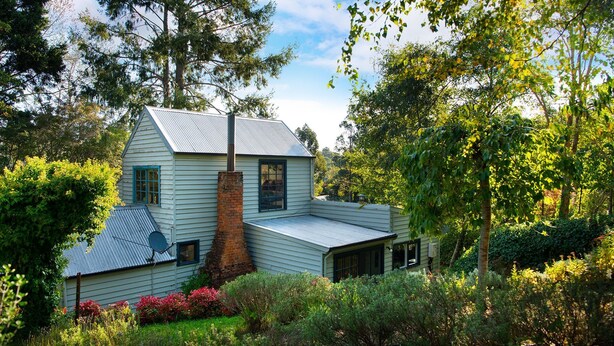Une maison à deux étages, dotée d’une cheminée et entourée d’une végétation luxuriante et d’arbres.