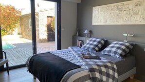 A bedroom with a bed, checkered bedding, a nightstand with a lamp, a bookshelf, and a view of the outdoors. - Calm'ré - Villa in a quiet area for family vacation (Sainte-Marie-de-Ré)