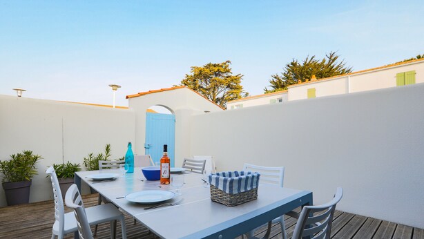 Une terrasse extérieure avec une table dressée pour le repas, entourée de chaises blanches et de plantes en pot.
