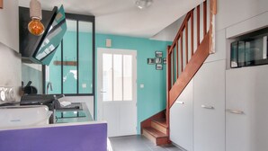 Une cuisine au design moderne, dotée d’un plan de travail blanc, d’un four encastré et d’un évier. Un escalier en bois mène à un palier où se trouvent une porte et une fenêtre.