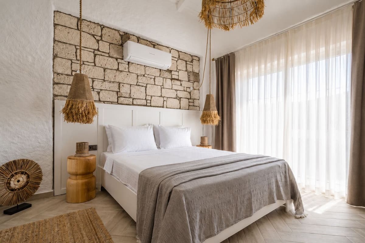 Un dormitorio con pared de piedra, una cama con ropa blanca, dos lámparas colgantes y un ventanal con cortinas.