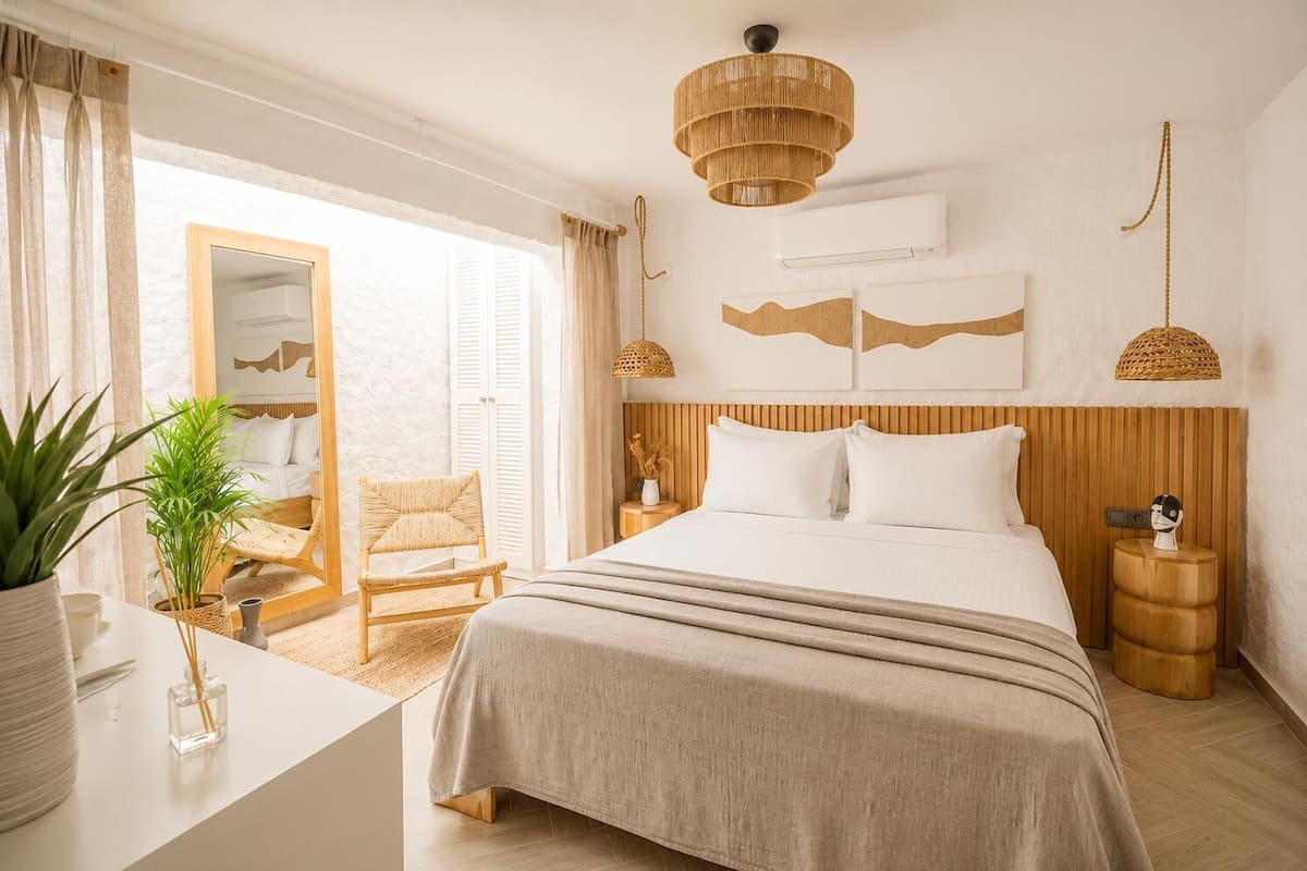 Un dormitorio con cama, una silla, una mesita de noche, una lámpara colgante y un espejo.