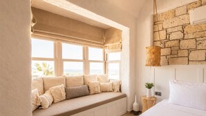 Une chambre avec un grand lit, un coin fenêtre agrémenté de coussins, une suspension décorative et une vue sur l’extérieur.