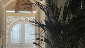 Un couloir avec une grande plante en pot, un luminaire suspendu en fibres tressées et une porte blanche dotée de panneaux de verre.