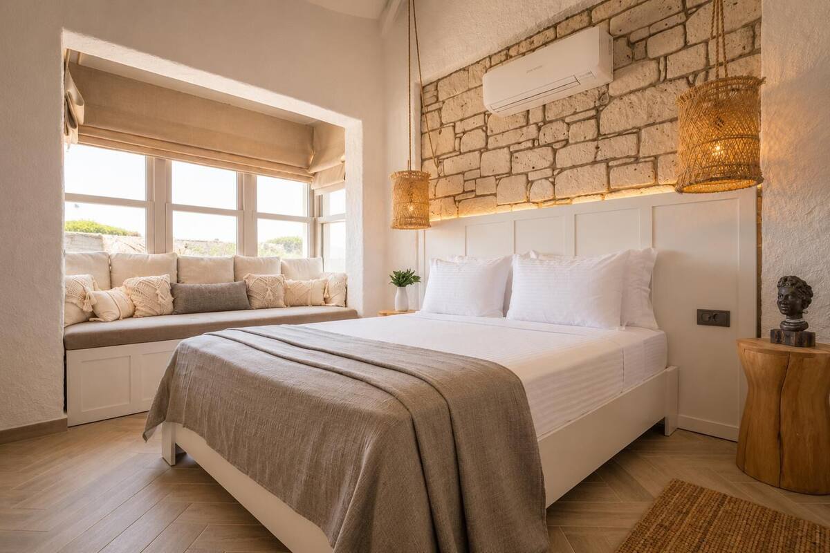Un dormitorio con cama, una banca junto a la ventana, pared de piedra y lámpara colgante.