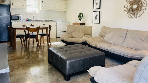 Ein modernes Wohnzimmer mit einem beigen Sofa, einem schwarzen Hocker und einem Essbereich mit Holztischen und -stühlen.