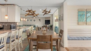 Un coin repas avec une table et des chaises en bois, une cuisine avec des meubles blancs et un salon avec un canapé et des œuvres d’art.