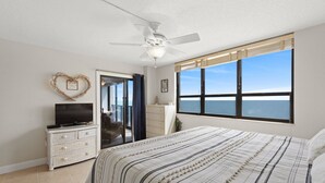 Ein Schlafzimmer mit Bett, Schminktisch, Fernseher, Deckenventilator und einem großen Fenster mit Meerblick.