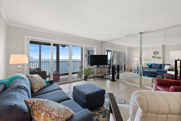 Un salón amplio con un sofá grande, una mesa de centro y un televisor. Hay un balcón con vistas al mar.