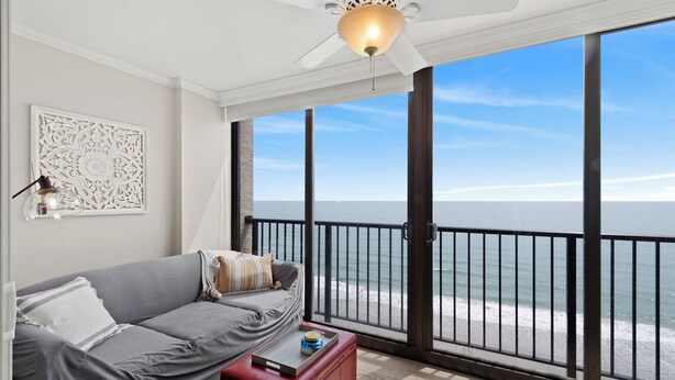 Ein Balkon mit Blick auf den Ozean, ein graues Sofa, ein roter Hocker und ein Deckenventilator.