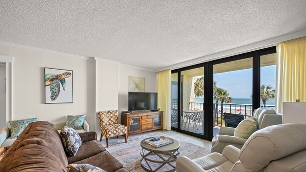 Una sala de estar con sofá, sillones, mesa de centro, televisor y balcón con vista a la playa.