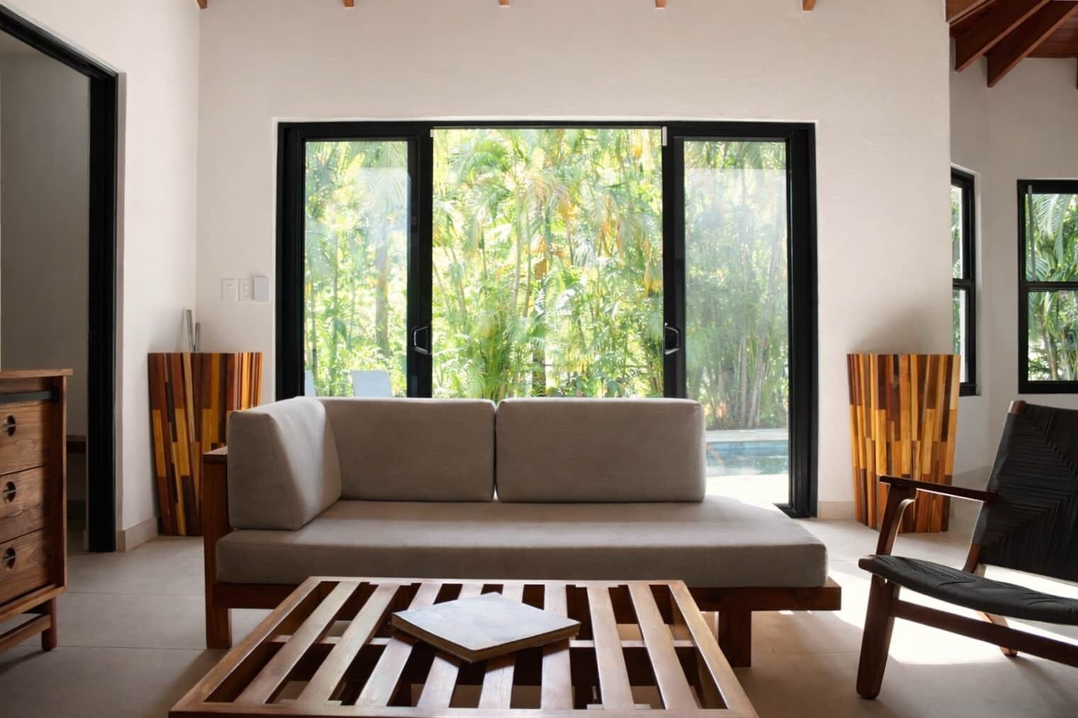 Ein modernes Wohnzimmer mit einer Couch, einem Holztisch und einem Sessel. Große Fenster bieten Blick auf üppiges Grün.