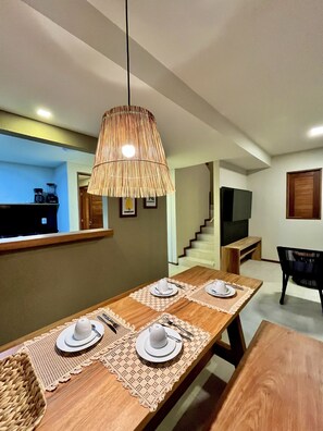 Un comedor con una mesa de madera dispuesta para cuatro personas, una lámpara colgante de bambú y una zona de cocina con cafetera y tostadora a la vista.