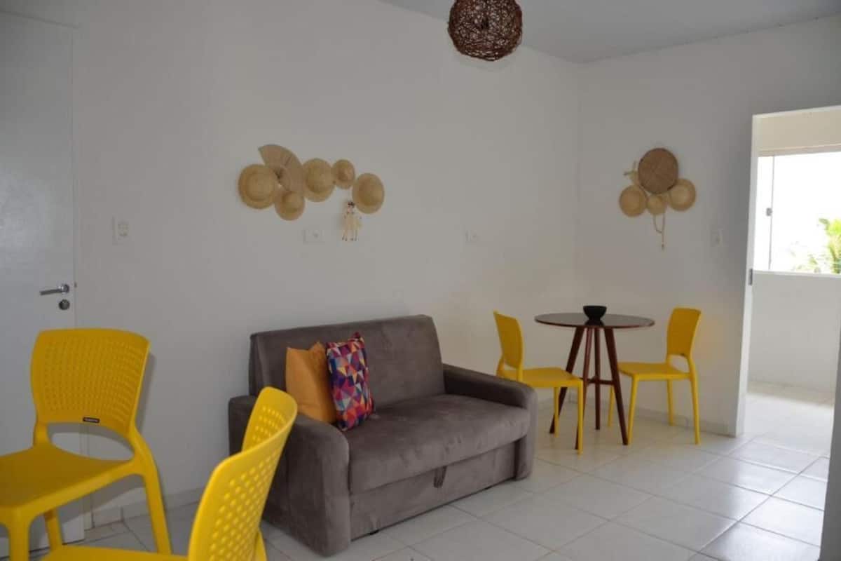 Een kamer met een grijze bank, gele stoelen, een kleine ronde tafel en wanddecoraties, waaronder hoeden en een hangmand.
