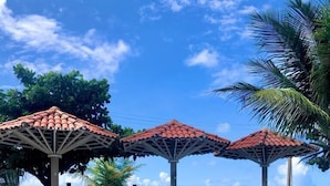 A swimming pool with tiled flooring, surrounded by lounge chairs and umbrellas, with a view of the ocean and palm trees. - Condominio Praias de Maragogi (Maragogi)