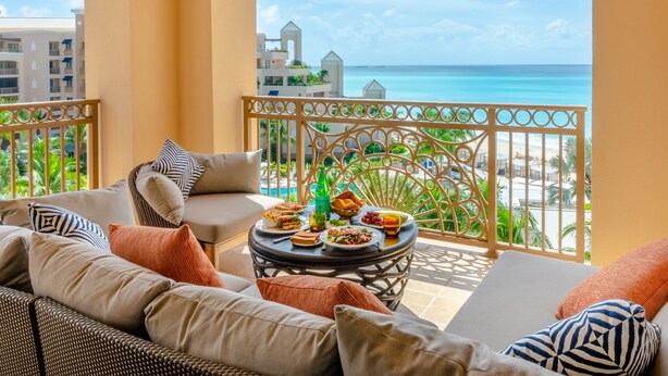 Varanda com vista para a praia, equipada com sofá, poltronas e uma mesa de jantar com comida.