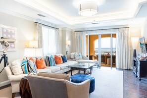 A spacious living room with a sofa set, a coffee table, and a television. There is a balcony with a view of the ocean.