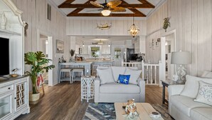 A spacious living room with a sofa, coffee table, and decorative items. A kitchen is visible in the background with a dining area. - The Beachcomber- Stunning Oceanfront Home in PKS, NC (Pine Knoll Shores)