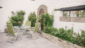 Un patio con una mesa redonda y cuatro sillas, rodeado por un muro blanco con una ventana de piedra y un muro de piedra con plantas.