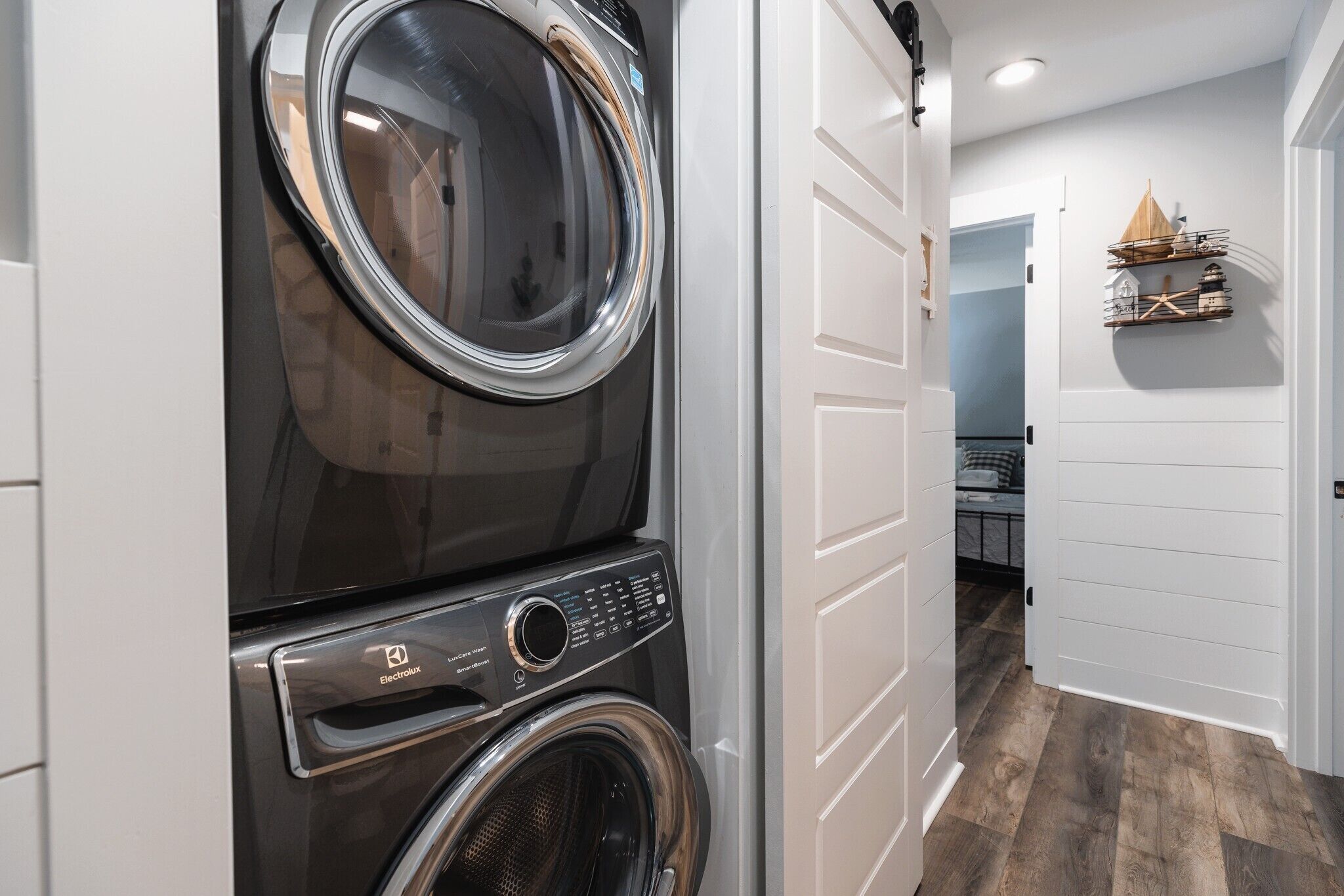 A laundry room with stacked washers and dryers, a white door, and a hallway leading to a bedroom.