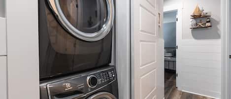 A laundry room with stacked washers and dryers, a white door, and a hallway leading to a bedroom.