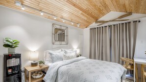 Une chambre moderne avec un plafond en bois, un lit avec une literie blanche, une table de chevet avec une lampe, un distributeur d’eau et une petite plante.