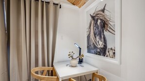 Une petite pièce avec une table blanche, deux chaises en bois, un vase de fleurs et un tableau représentant un cheval, encadré, accroché au mur.