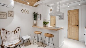 Une cuisine moderne avec un plafond en bois, des murs blancs et une porte en bois. On y trouve deux tabourets, une petite table et un luminaire suspendu.