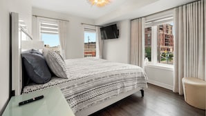 A bedroom with a bed, a TV mounted on the wall, a window with curtains, and a view of buildings outside. - Etobicoke Suites (Toronto)