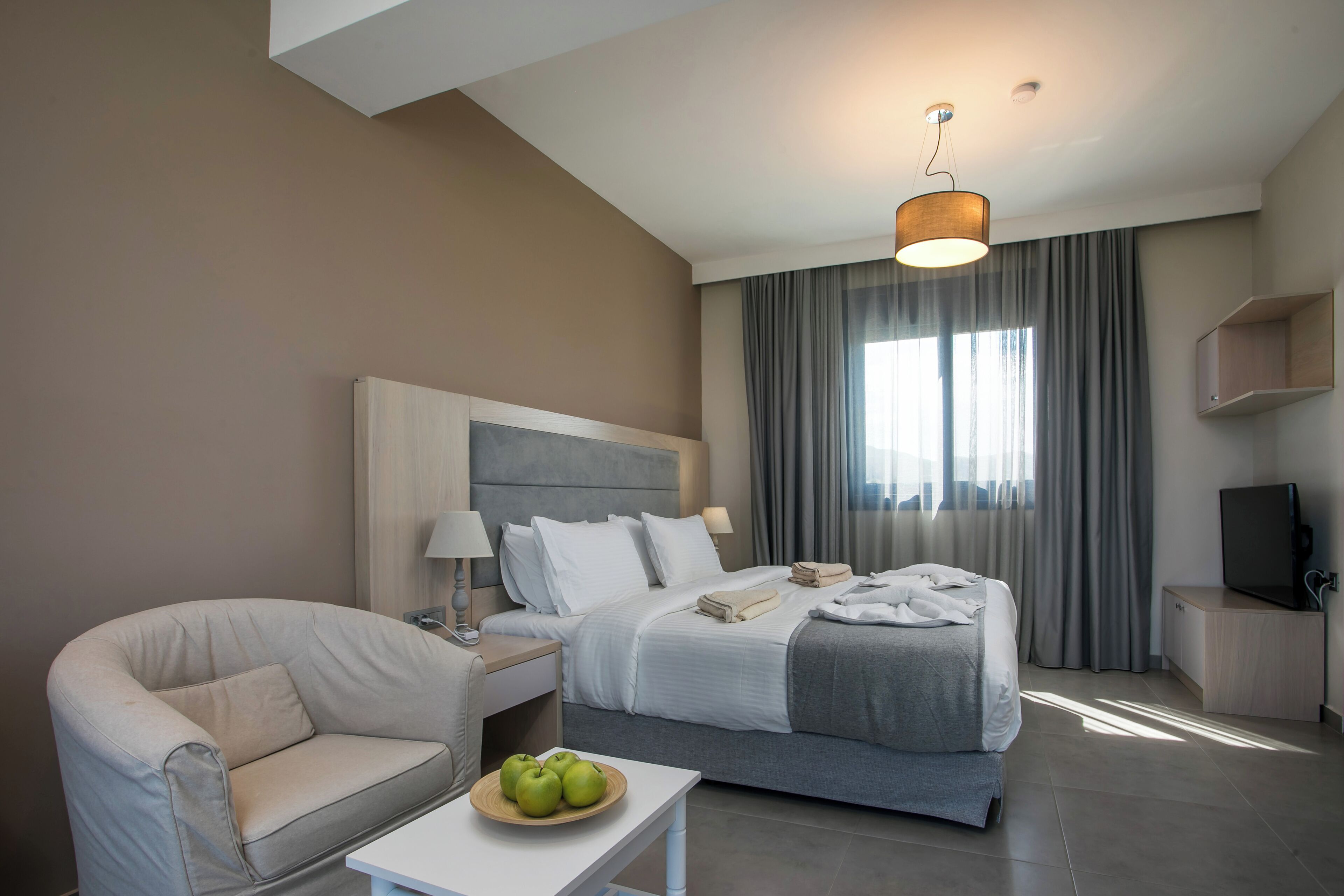 A modern hotel room with a bed, a sofa, a small table with apples, a television, and a large window with curtains.