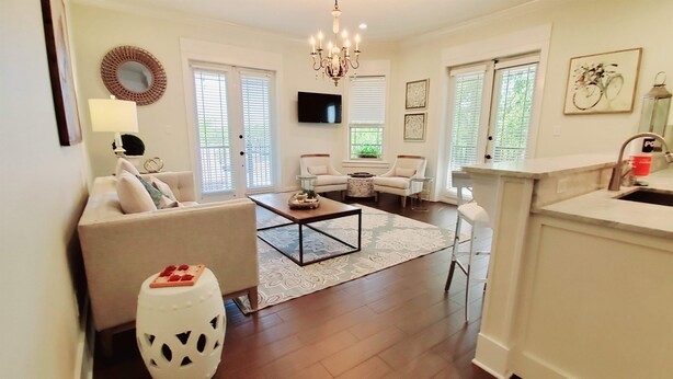 A spacious living room with a sofa, coffee table, and a TV. There are two windows with blinds, a chandelier, and a kitchen area with a sink.