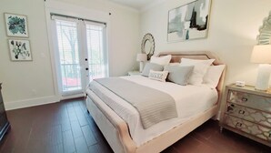 A bedroom with a large bed, a nightstand with a lamp, and a dresser. There are two windows with blinds, and artwork on the walls.