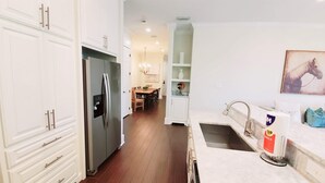 A modern kitchen with white cabinetry, a stainless steel refrigerator, a sink with a towel dispenser, and a dining area in the background.