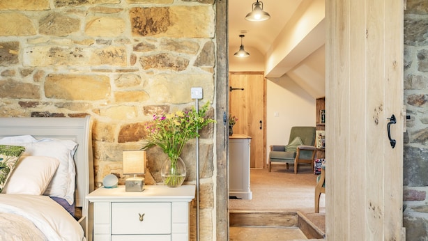 Une chambre avec un mur en pierre, une porte en bois, une table de chevet avec une lampe et un lit avec des oreillers.