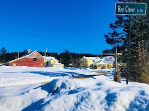 Red Clover Inn in einer verschneiten Landschaft, mit einem roten Bauernhaus und gelben Häusern.