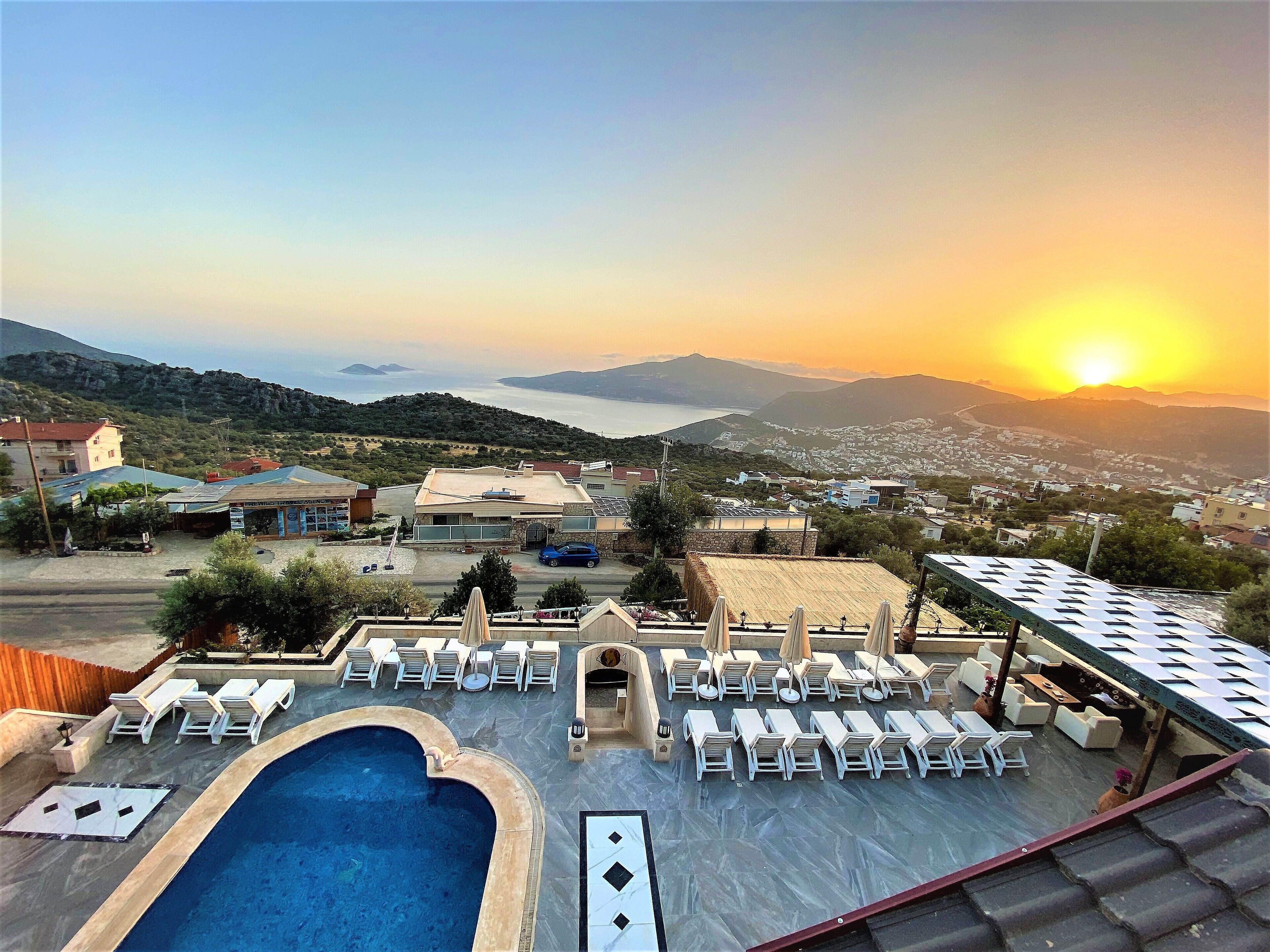 Un área de piscina en la azotea con sillones de descanso y una piscina, con vistas a un pueblo costero y montañas durante el atardecer.