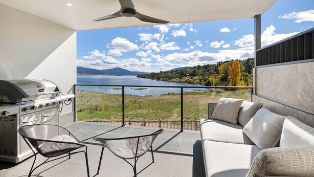A balcony with a grill, two chairs, and a sofa overlooking a lake and mountains. - Lake Villavista - 4BR Luxury Townhouse l Pet Friendly l Lake Views l WiFi l BBQ (East Jindabyne)