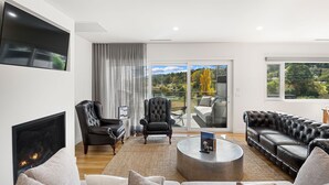 A modern living room with a fireplace, leather sofas, and a large window overlooking a scenic landscape. - Lake Villavista - 4BR Luxury Townhouse l Pet Friendly l Lake Views l WiFi l BBQ (East Jindabyne)