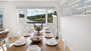 A dining room with a wooden table set for a meal, a view of a lake and hills through large windows, and a modern lamp. - Lake Villavista - 4BR Luxury Townhouse l Pet Friendly l Lake Views l WiFi l BBQ (East Jindabyne)