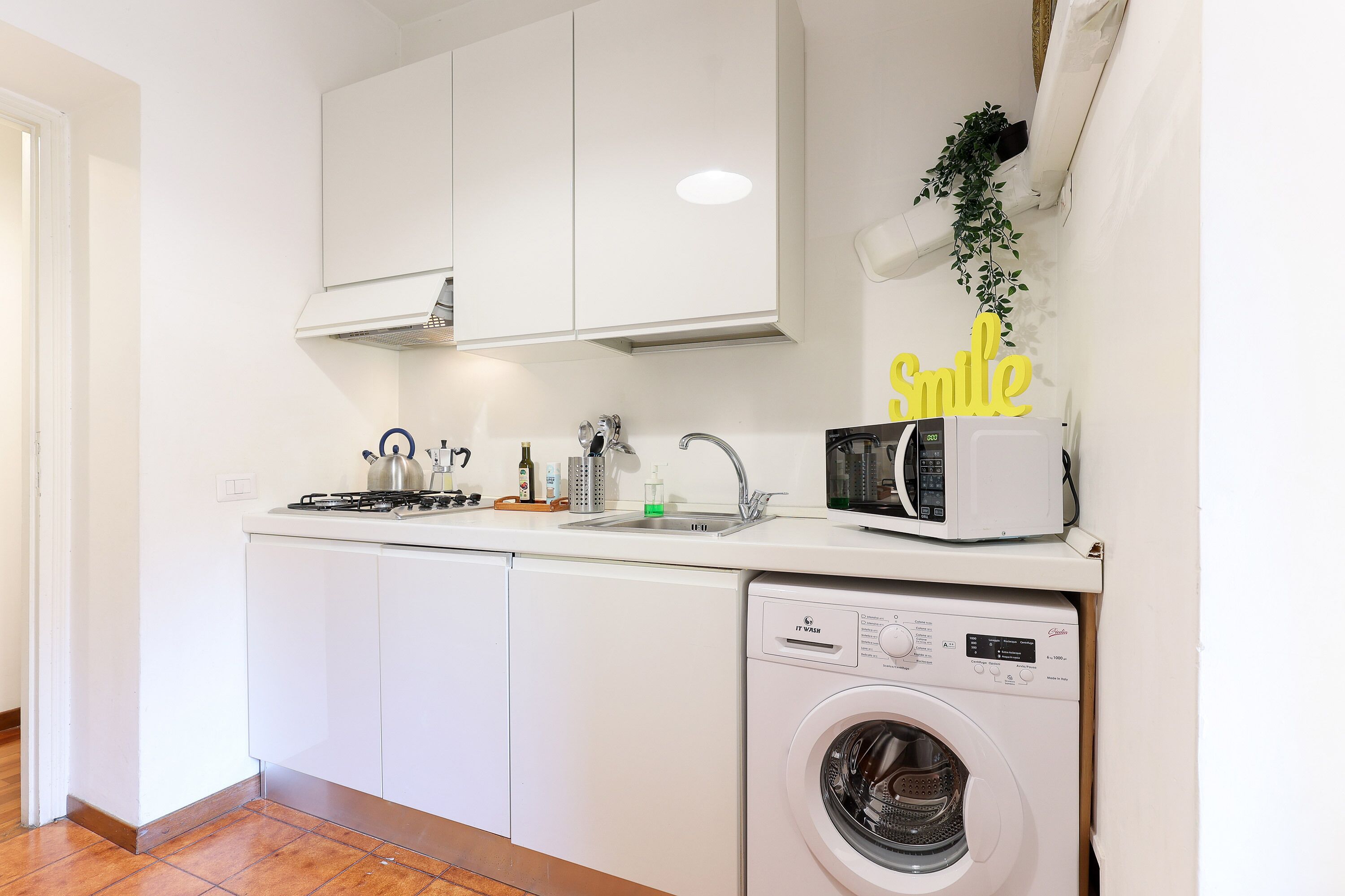 Una cocina compacta con armarios blancos, microondas, lavamanos y lavadora.