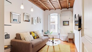 Un salon chaleureux avec un canapé marron, une table basse ronde sur laquelle se trouvent une bouteille et des verres, et une fenêtre donnant sur des immeubles.