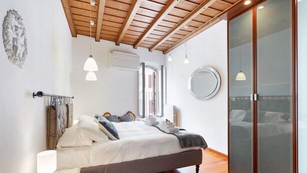 Une chambre avec un plafond en bois, un lit recouvert de linge de lit blanc, une lampe de chevet, un miroir et une armoire aux portes en verre dépoli.